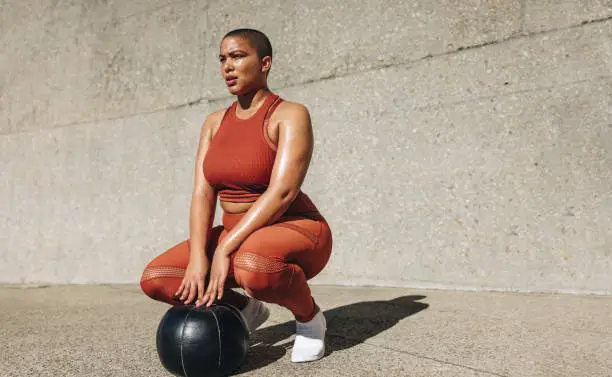 Fitness woman crouching with medicine ball on ground. Plus size female in sportswear with medicine ball looking away.