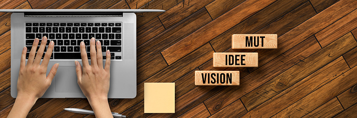cubes with German message for VISION, IDEA, COURAGE and hands over laptop on wooden background