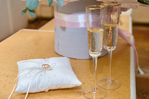Close up of two golden wedding rings on pillow, sparkling glasses with champagne and bouquet of flowers in hat box on table, copy space. Wedding concept