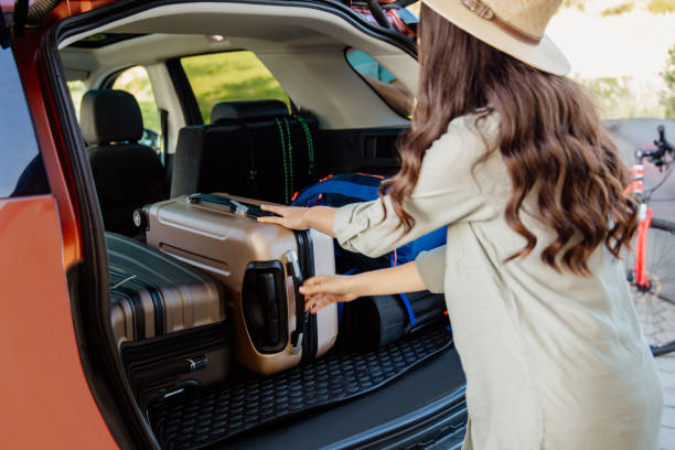 Woman holding pulling luggage in car trubk Rear view of a woman holding bag standing near car boot trunk stock pictures, royalty-free photos & images