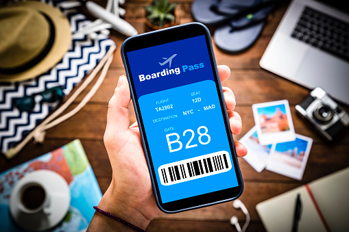 Top view of a caucasian hand holding a smartphone with a electronic boarding pass on it screen and surrounded by some defocused traveling stuff such as a camera, snapshots, a map, a laptop, and some summer clothing. The smartphone screen is at the center of the image. Studio shot taken with Canon EOS 6D Mark II and Canon EF 24-105 mm f/4L