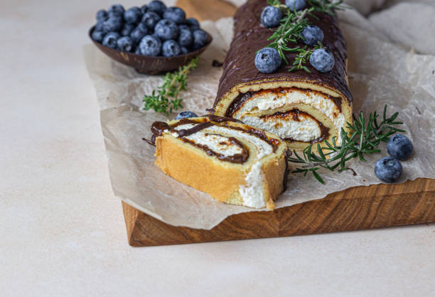 Sponge cake roll with chocolate and cream cheese decorated with chocolate glaze, blueberry and rosemary on parchment, light background. Biscuit swiss roll.