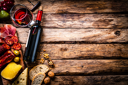 Appetizer: top view of a rustic wooden table with a composition of red wine bottle, wineglass, a cutting board with various cheeses and Iberico ham arranged at the left border leaving useful copy space for text and/or logo at the right. Some grapes, olives, nuts, bread and a corkscrew complete the composition. Predominant colors are red and brown. High resolution 42Mp studio digital capture taken with SONY A7rII and Zeiss Batis 40mm F2.0 CF lens