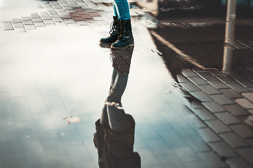 A woman in boots stands near the puddles. Reflections of the sky in puddles. Lower angle. The concept of psychology and loneliness.