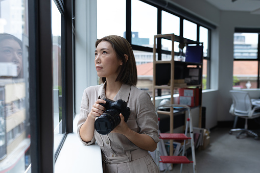 Asian businesswoman holding camera looking through window in creative office. social distancing in workplace during covid 19 pandemic.