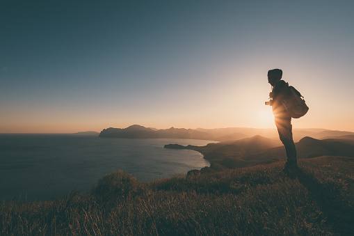 Young standing man with backpack. Hiker on the stone on the seashore at colorful sunset sky. Beautiful landscape with sporty man rocks sea and clouds at sunset. Sporty lifestyle