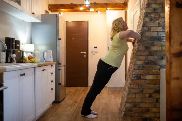 Mature woman doing standing wall push-ups at home during coronavirus pandemic.