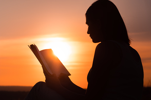 silhouette of a young beautiful woman at dawn sitting on the ground and carefully staring at the open book