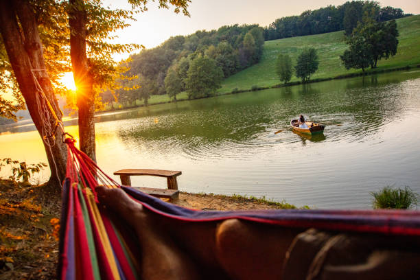 uomo che si rilassa sull'amaca mentre la donna rema una barca in canoa sul lago - canoeing people traveling camping couple foto e immagini stock