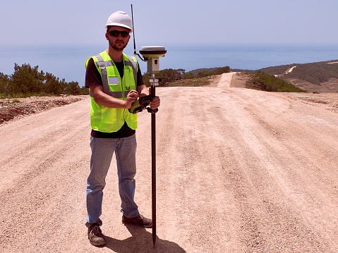 Geodetic engineer surveyor in white hard hat doing measurements with GNSS satellite receiver during road construction works.