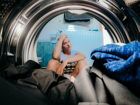 Unhappy woman sitting with head in hands in front of washing machine at bathroom