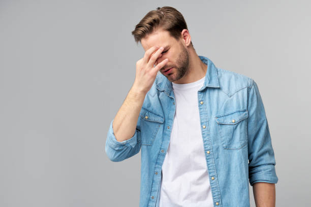 istock Frustrated young caucasian man in jeans shirt touching head with hand while standing overg studio grey background 1297420815