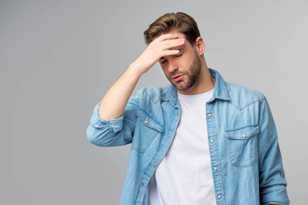 istock Frustrated young caucasian man in jeans shirt touching head with hand while standing overg studio grey background 1297420817