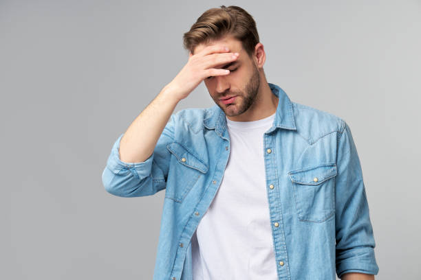 istock Frustrated young caucasian man in jeans shirt touching head with hand while standing overg studio grey background 1297420818