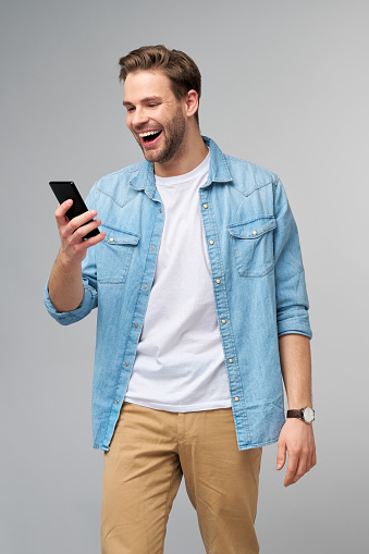 Smiling young man wearing jeans shirt taking selfie photo on smartphone or making video call standing over grey studio background.
