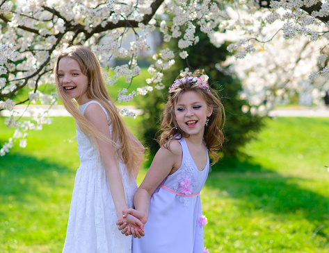 Two beautiful young girls with blue eyes in a white dresses in the garden having fun and enjoying smell of flowering spring garden.