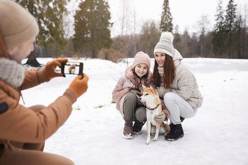 Portrait of father taking photo of cute daughter and wife with dog while enjoying walk outdoors together in winter forest, copy space