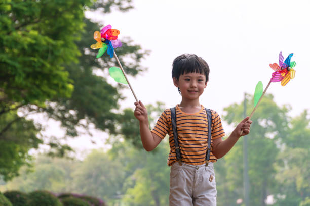 istock Happy little boy holding windmill in his hands while standing with tree in morning at the public park. Child cute boy playing pinwheel while walking find windy at garden park. 1297426008