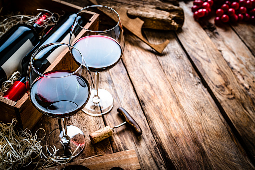 High angle view of two red wineglasses shot on rustic wooden table. A wine bottle box is beside the wineglasses. Selective focus on foreground.  A corkscrew and grapes complete the composition. The composition is at the left of an horizontal frame leaving useful copy space for text and/or logo at the right. Predominant colors are red and brown. High resolution 42Mp studio digital capture taken with SONY A7rII and Zeiss Batis 40mm F2.0 CF lens