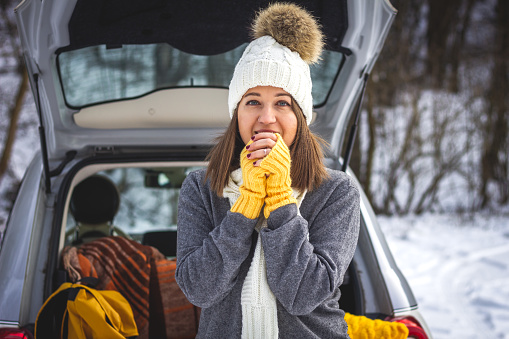 Woman with knit hat warming up herself next to car during travel to winter vacation