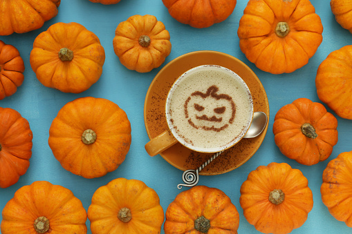 Stock photo showing elevated view of autumnal, Halloween scene of orange, coffee cup on saucer with metal tea spoon surrounded by gourds arranged on turquoise blue background. The cup contains a hot Latte decorated with a Jack O'Lantern design latte milk foam art.