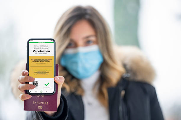 A defocused young girl, wearing a face mask, is holding a passport and a smart phone with a with an example of a certificate of vaccination against the Covid-19 disease. (Selective focus, focus on the smart phone) A defocused young girl, wearing a face mask, is holding a passport and a smart phone with a with an example of a certificate of vaccination against the Covid-19 disease. immunization certificate photos stock pictures, royalty-free photos & images