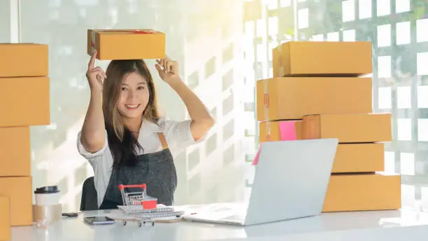 Photo of Young Asian working at home and starting a small business SME with online internet. And a laptop on the table, a girl holding a box on her head