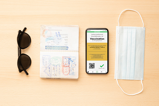 A face mask, passport, sunglasses and a smart phone with an example of a certificate of vaccination against the Covid-19 disease, are arranged on a wooden table.