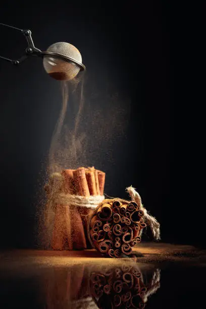 Cinnamon powder is poured out of the strainer. The ground cinnamon, cinnamon sticks, tied with jute rope on a black reflective background.  Copy space.