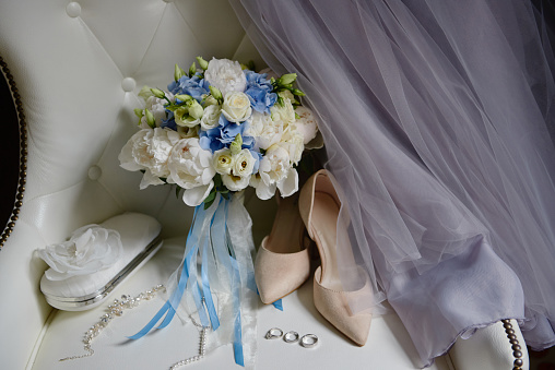 Bridal bouquet of white and blue flowers with satin ribbon, beige women shoes, two golden wedding rings and purple dress on armchair, copy space. Wedding concept