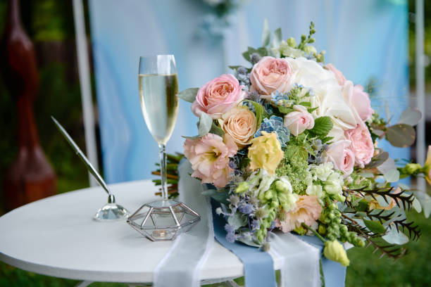 Close up of bridal bouquet of pink and blue flowers, glass of champagne and wedding rings on white wood table outdoors, copy space. Wedding concept stock photo
