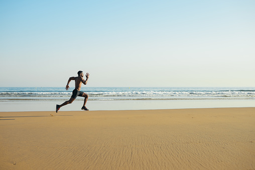 Black fit man running fast by the sea on the beach. Powerful runner training outdoor on summer.