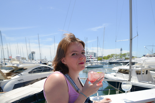 Close up shot of pretty young woman enjoying a cool drink at the harbour with yachts in the background.