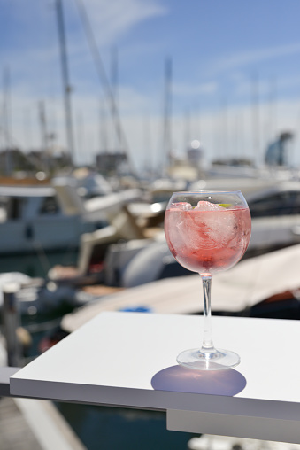 Glass of  alcohol rests on a table in the sunshine with expensive yachts in the background in the harbour at Barcelona.