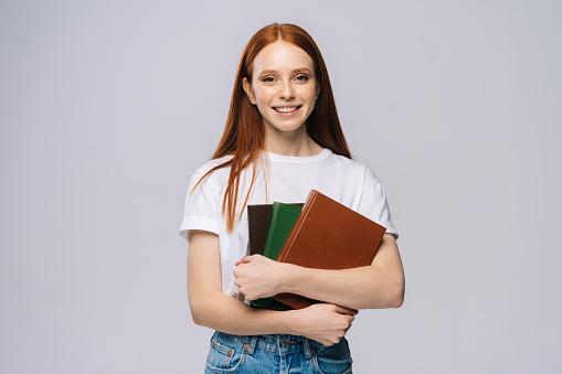 Smiling happy young woman college student wearing T-shirt and denim pants holding book on isolated gray background. Pretty redhead lady model emotionally showing facial expressions, copy space.