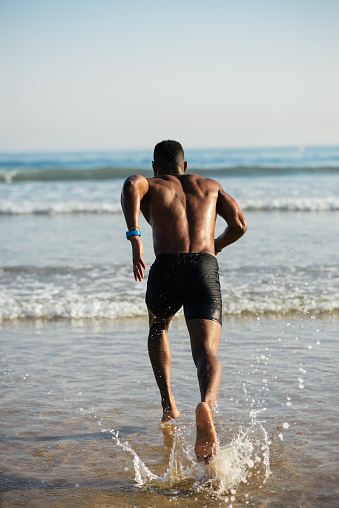 Athlete running to the sea for swimming sport training. Back view of black swimmer splashing water.