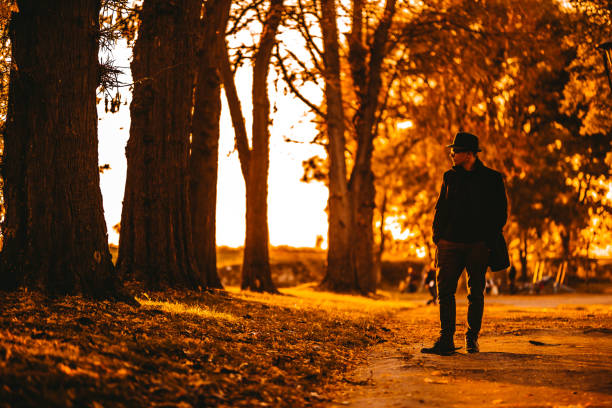 Portrait of a handsome young male model wearing black hat and long black coat, he is walking in autumn public park.