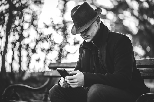 Portrait of a handsome young male model wearing black hat and long black coat, he is sitting on bench in autumn public park and checking social media or emails on smartphone.