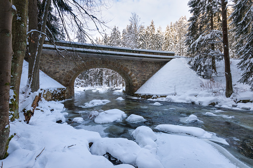 Wild river in winter with old stone bridge at Zemska Brana nature reserve, Orlicke hory, Eagle mountains, Eastern Bohemia Czech republic. Beautiful frosty day. Snowy weather in mountain. Most popular