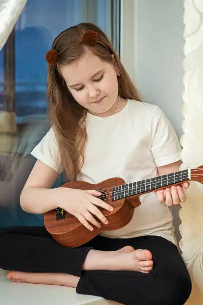 Photo of Little cute girl in white t-shirt and black leggings sitting inside of home and playing ukulele.