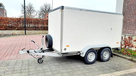 White vehicle trailer closed model in good condition with four wheels and a spare wheel. Trailer on an empty parking place on grey tiles. Liw beach trees hedge en some trees in the background