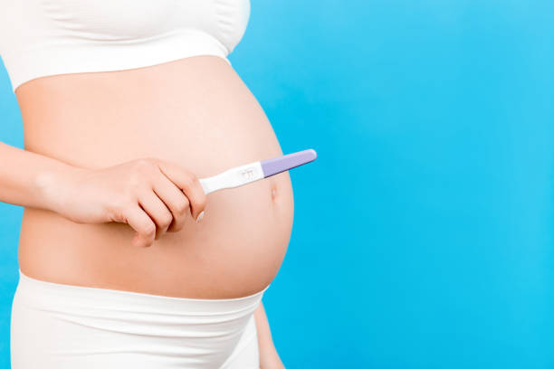 Close up of positive pregnancy test against pregnant woman's belly at blue background. Future mother in white underwear. Baby expecting. Copy space Close up of positive pregnancy test against pregnant woman's belly at blue background. Future mother in white underwear. Baby expecting. Copy space. babyproof stock pictures, royalty-free photos & images