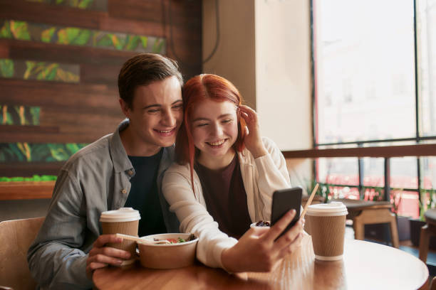 istock Happy teenage couple smiling at camera while taking selfies using smartphone, sitting in a cafe together on a daytime 1297486265