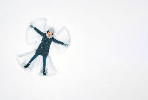 Young beautiful woman in a gray jacket, hat and gloves, blue jeans, boots makes a snow angel in the snow, a view from a drone. Copy space, banner. Snow fun, entertainment