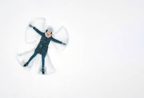 Young beautiful woman in a gray jacket, hat and gloves, blue jeans, boots makes a snow angel in the snow, a view from a drone. Copy space, banner. Snow fun, entertainment Young beautiful woman in a gray jacket, hat and gloves, blue jeans, boots makes a snow angel in the snow, a view from a drone. Copy space, banner. Snow fun, entertainment snow angels stock pictures, royalty-free photos & images