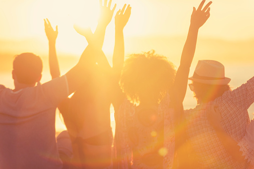Group of friends sitting on the beach at sunset/sunrise. They are watching the sun set over the ocean. They are celebrating with their arms in the air. Backlit silhouette with copy space.