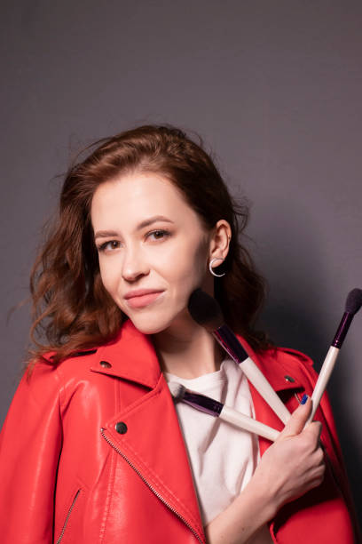 istock charming brunette woman in red leather jacket with makeup brushes isolated on grey wall background. professional makeup artist 1297489487