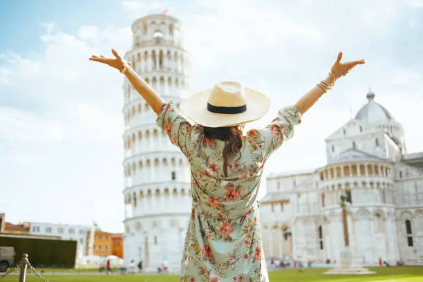 Seen from behind tourist woman in floral dress with hat near Leaning Tower in Pisa, Italy with open arms rejoicing.