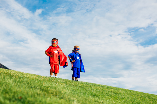 Two young boys, dressed up as superheroes, and ready to conquer their fears and show their strength in life. They are standing in a public park during summer.