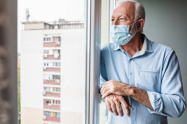 istock Worried senior man with face mask standing by the window 1297499075